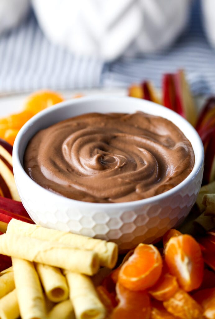 A close up of a white bowl full of vegan chocolate dip that's surrounded by orange slices, cookies, and apple slices.