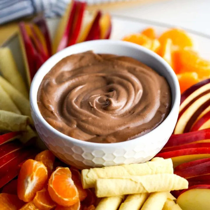 Creamy vegan cashew chocolate dip in a white bowl surrounded by fruit and rolled cookies.