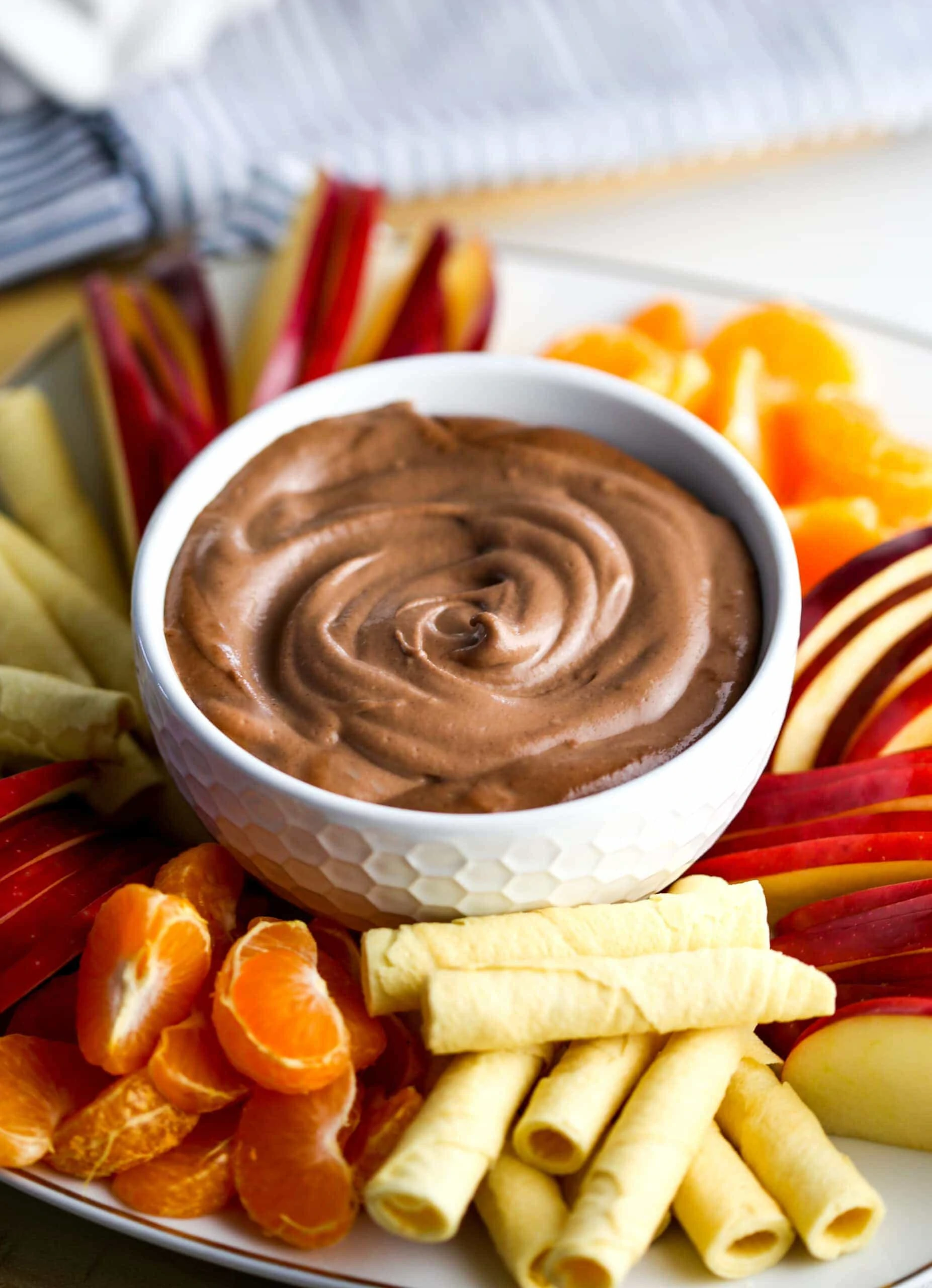 Creamy vegan cashew chocolate dip in a white bowl surrounded by fruit and rolled cookies.
