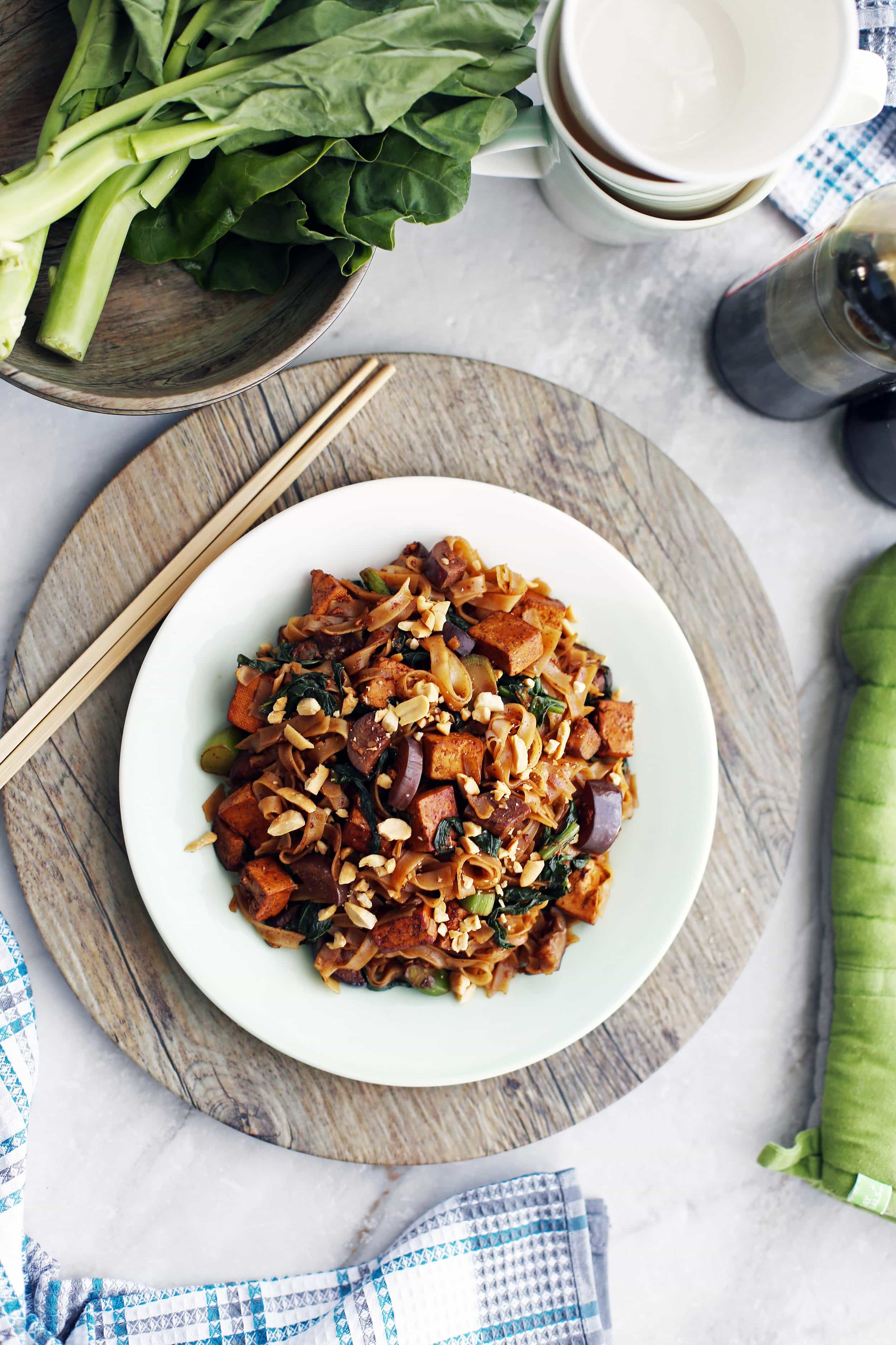 Vegetarian pad see ew with tofu and Chinese eggplant on a white plate; chopsticks to its side.