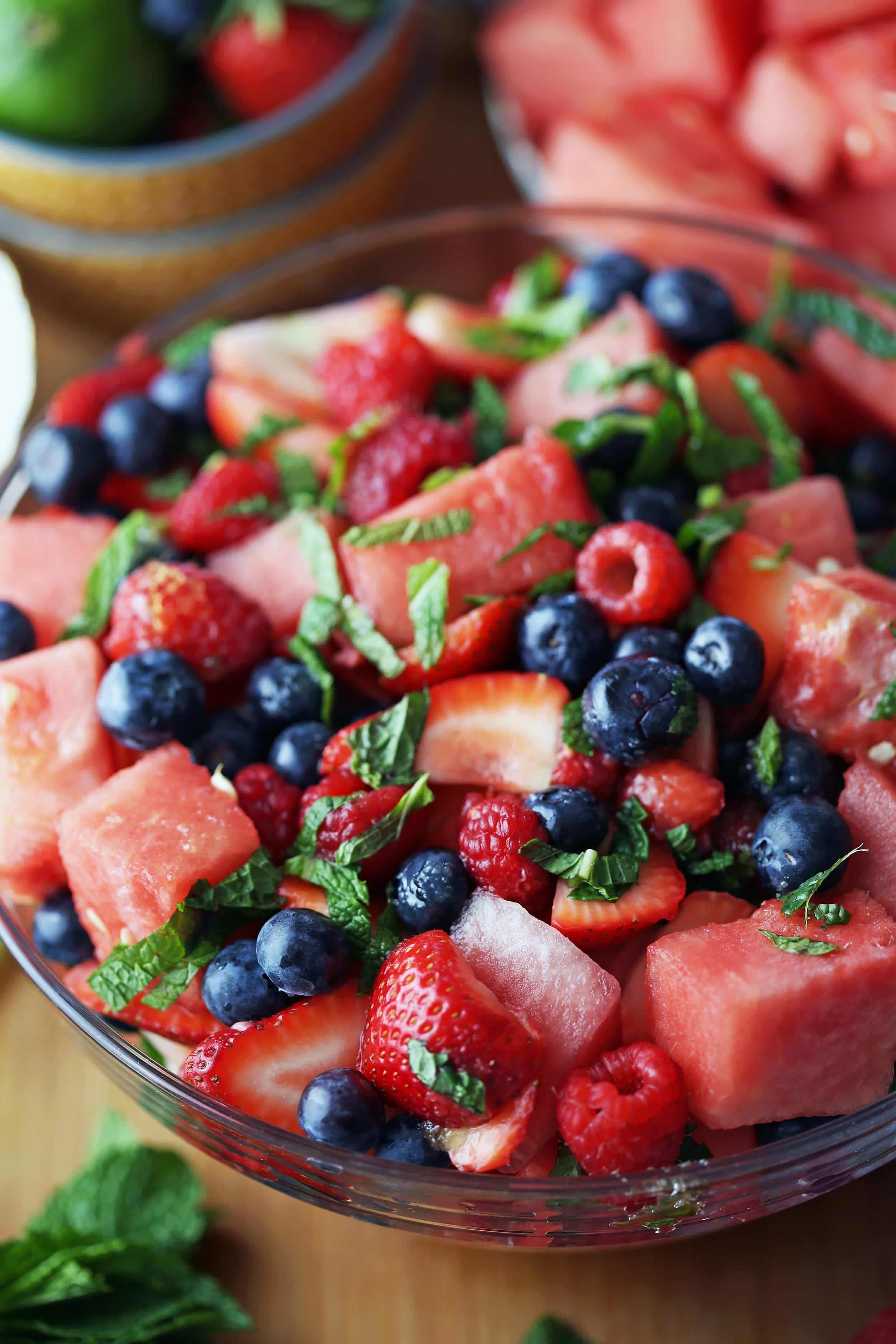 A glass bowl holding a fruit salad, which includes watermelon. strawberries, blueberries, and raspberries.