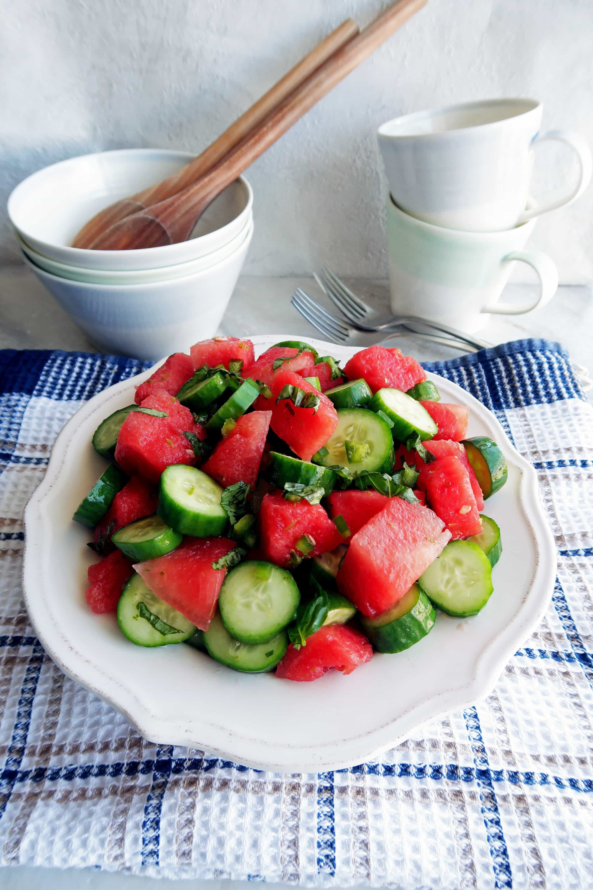 Watermelon Cucumber Jalapeno Salad on a white plate with three bowls, two wooden spoons, two forks, and two cups in the background.
