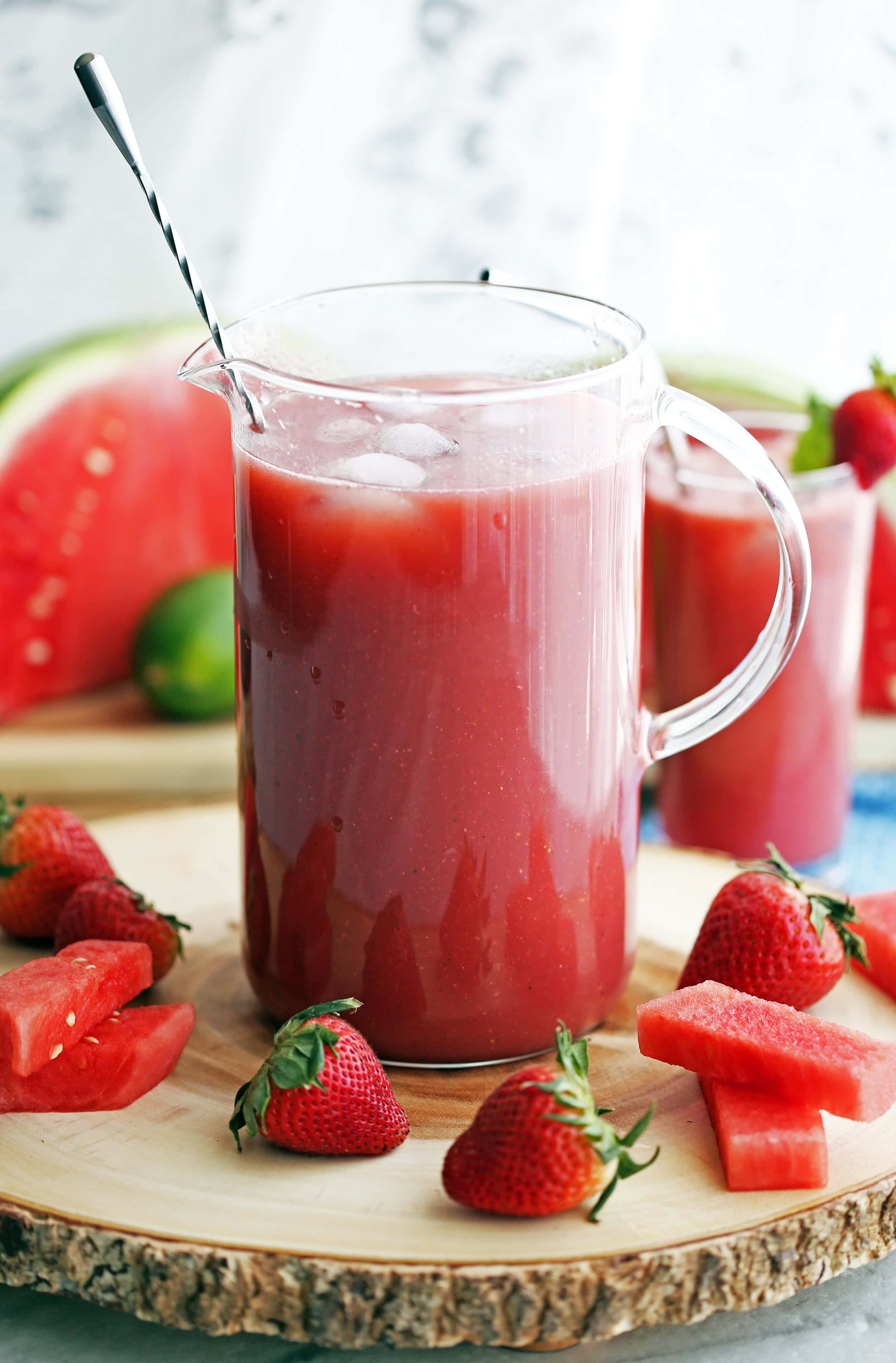 A glass pitcher full of Fresh Watermelon Strawberry Soda that's surrounded by fresh strawberries and watermelon pieces.