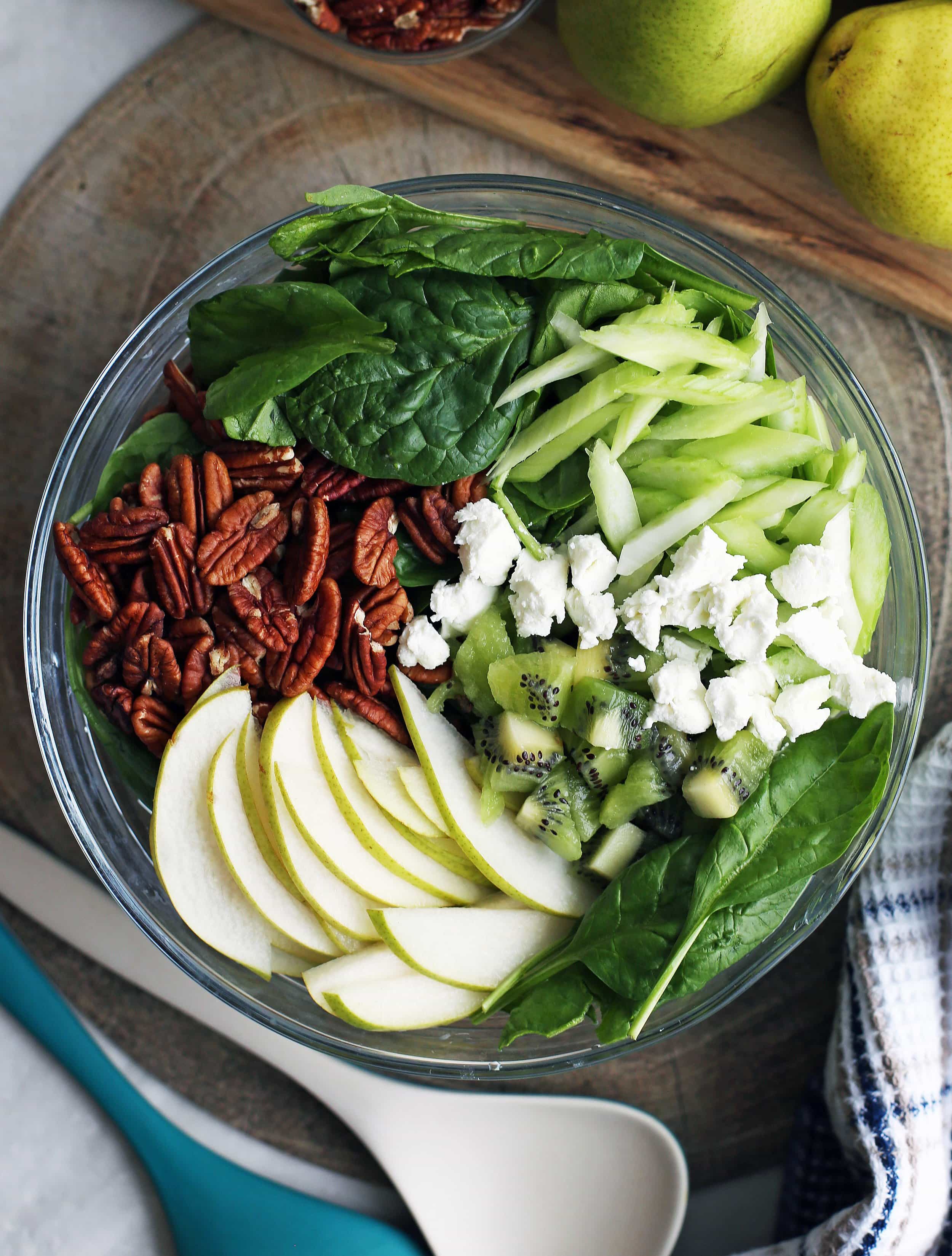 Winter Green Salad containing spinach, pears, kiwi fruit, goat cheese, and pecans in a large glass bowl.