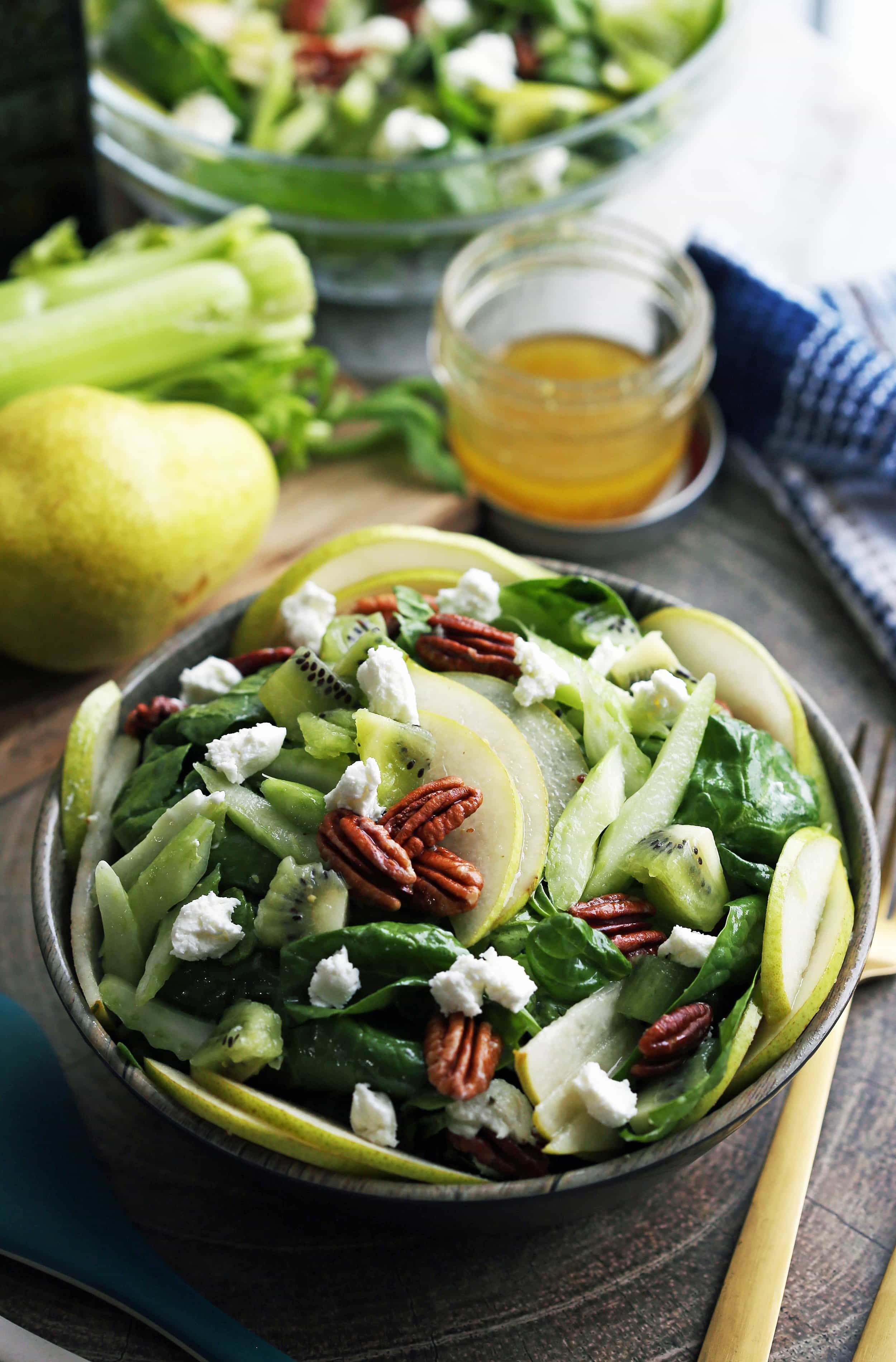 A bowl full of winter green spinach salad with pears, kiwi, and pecans with vinaigrette in a small mason jar to its side.