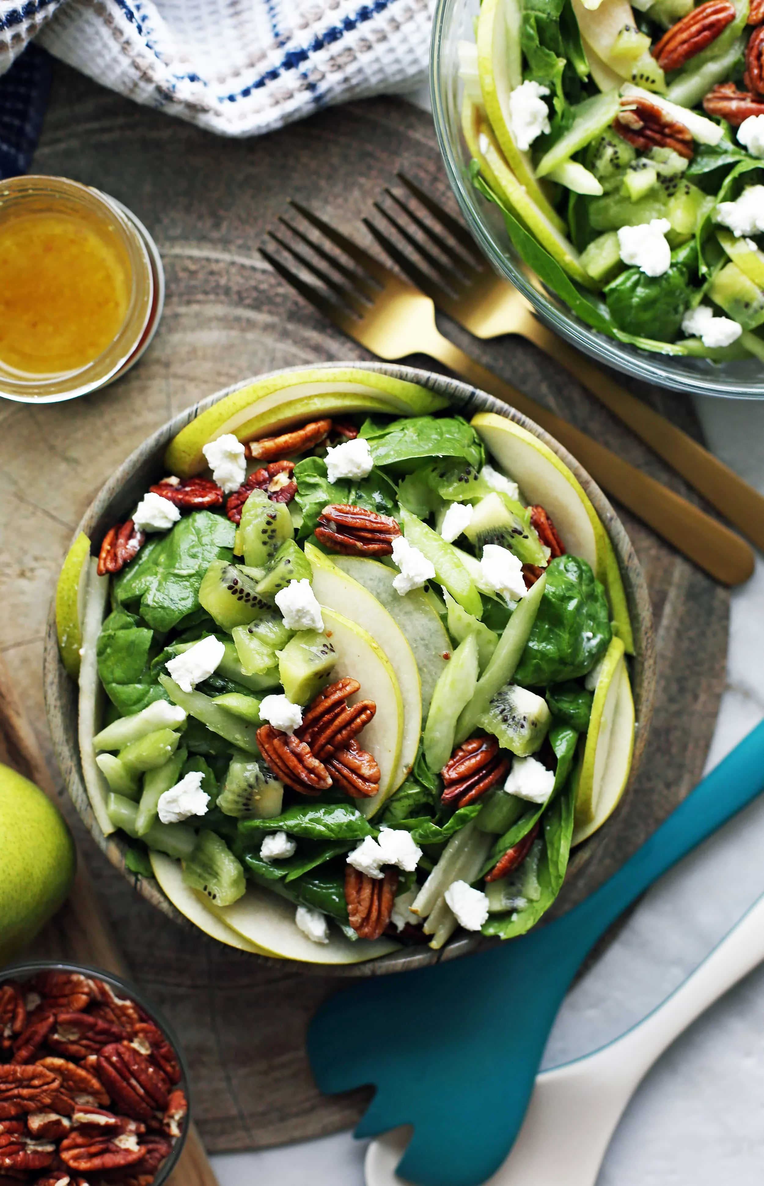 Overhead view of a wooden bowl of winter green spinach salad with bowls of more salad, pecans, and vinaigrette surrounding the bowl.