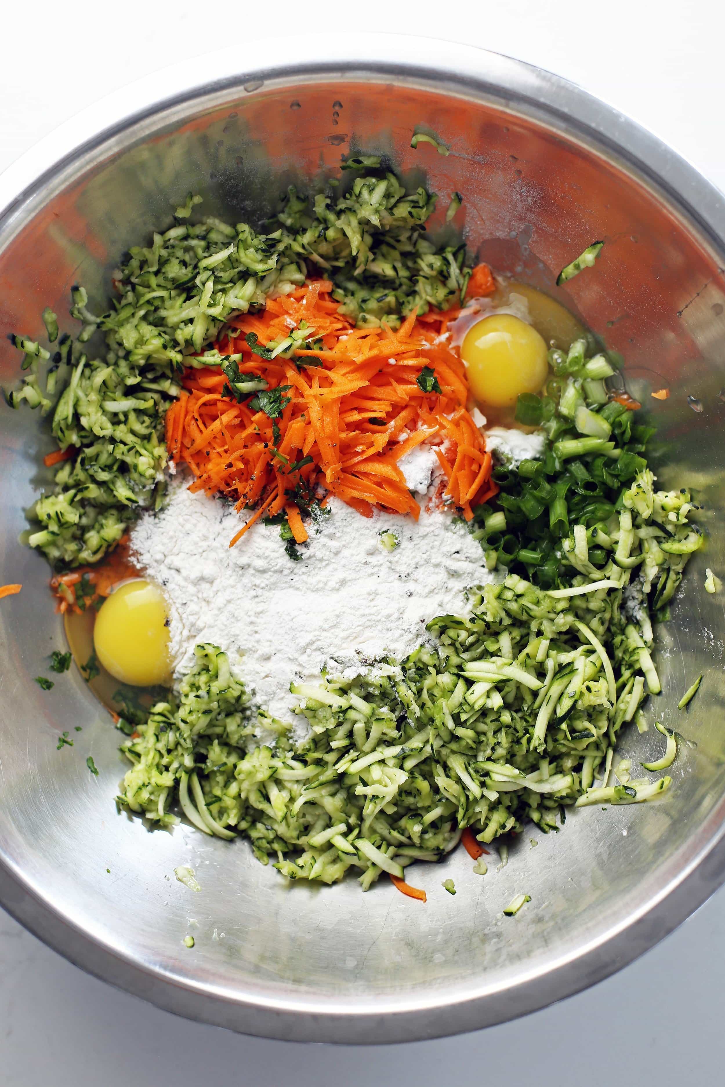 Shredded zucchini, shredded carrots, green onion, parsley, eggs, flour, baking powder, and cracked black pepper in a large stainless steel bowl.
