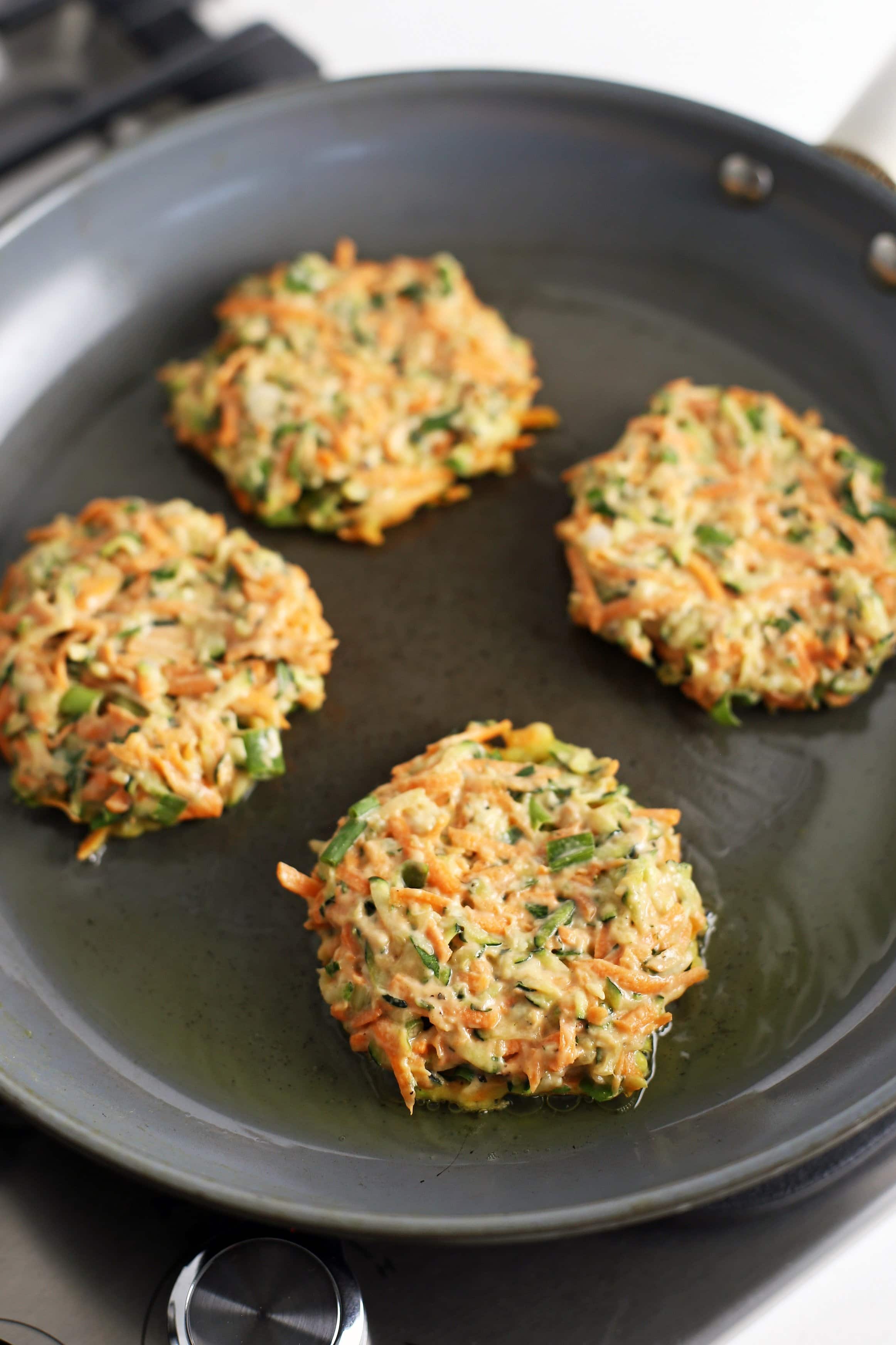 Four zucchini carrot pancakes frying in a small amount of olive oil in a large frying pan.