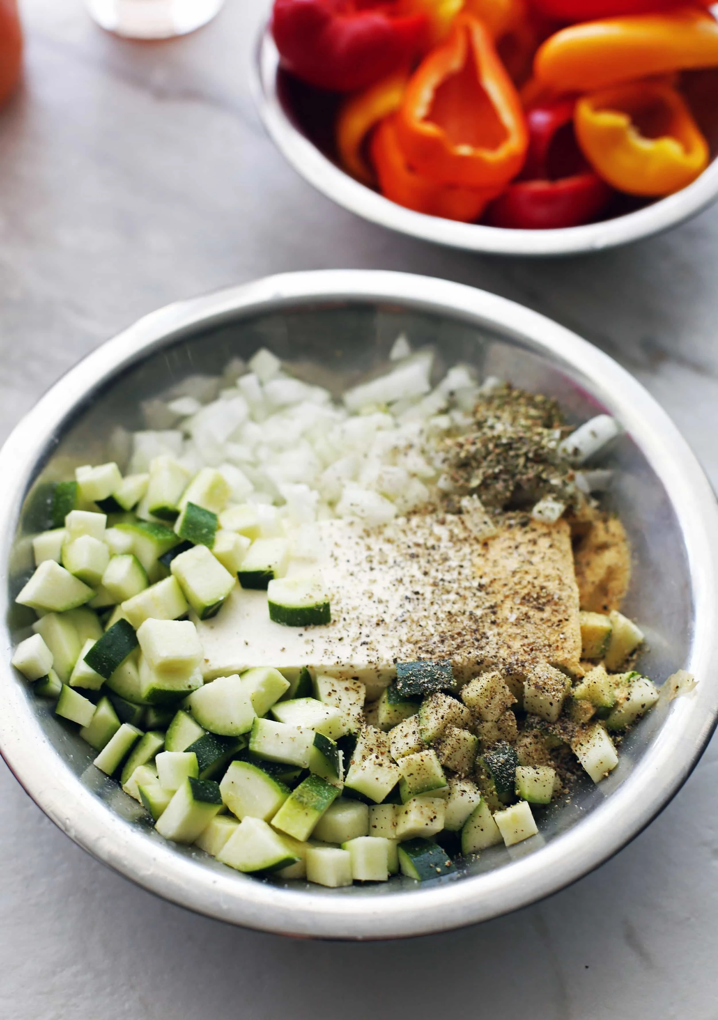 A metal bowl containing cream cheese, diced zucchini, onion, dried herbs, and spices.